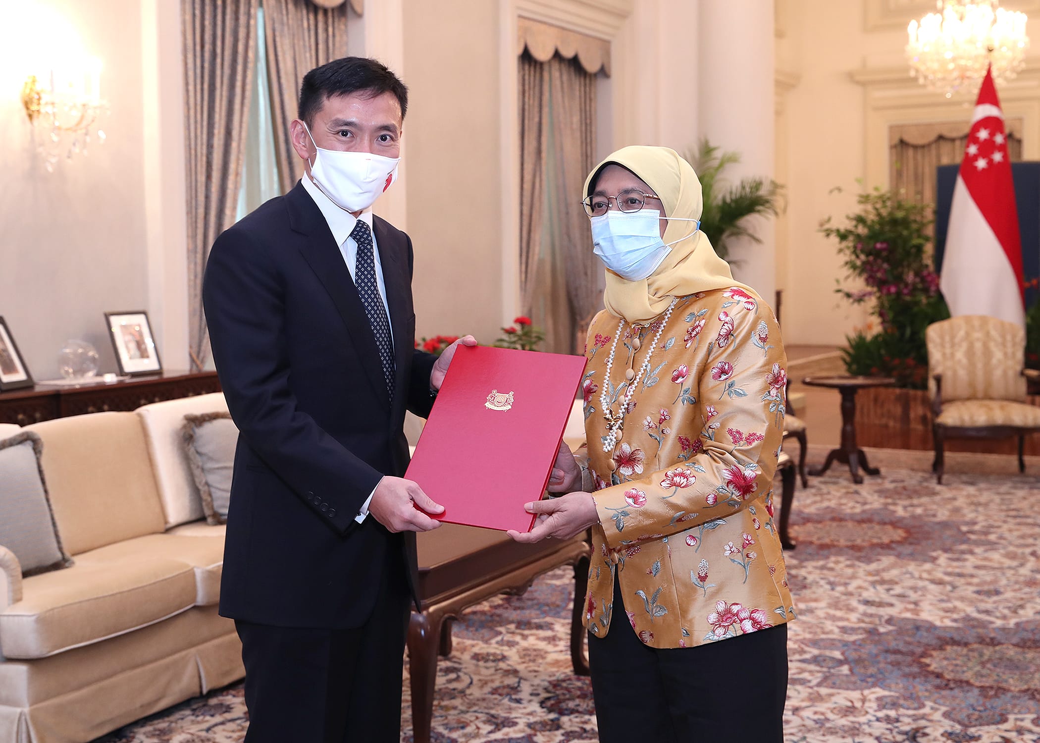 Two people wearing masks exchanging a red folder with a crest in an ornate room.